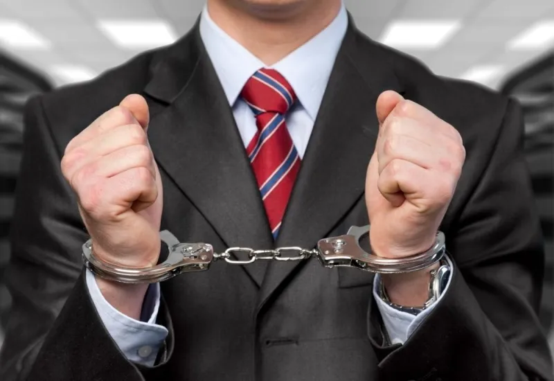 Man in a dark suit and striped red tie with his wrists in handcuffs, face out of frame.