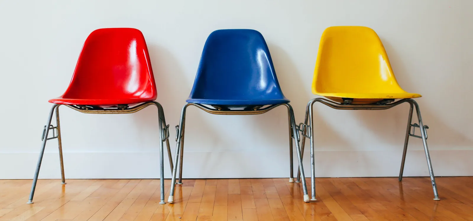 Three chairs in red, blue and yellow lined up against a white wall.