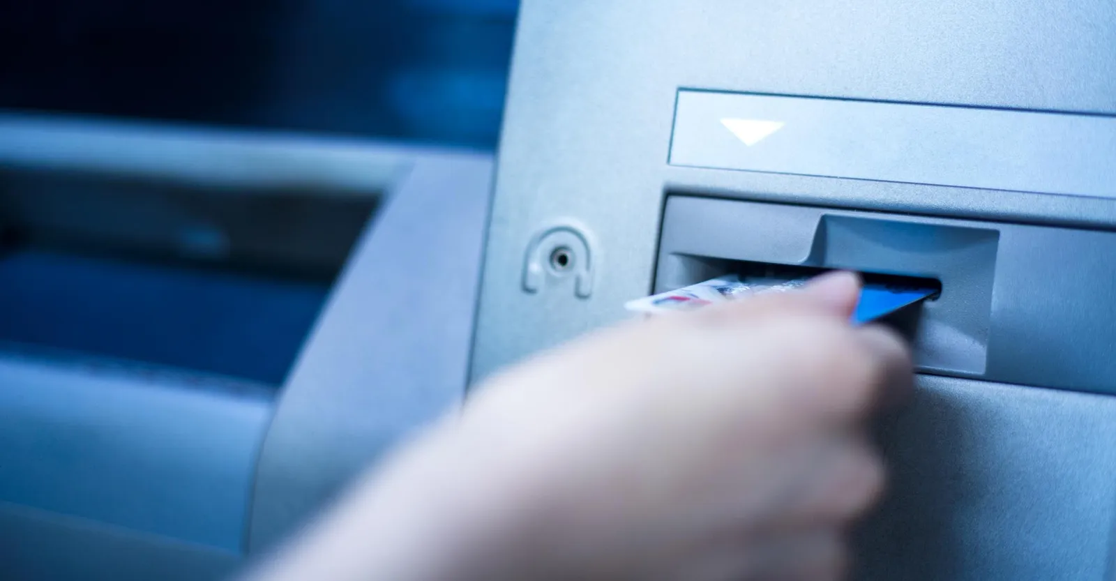 Close-up of a hand inserting a card into a cash machine, in blue tones.