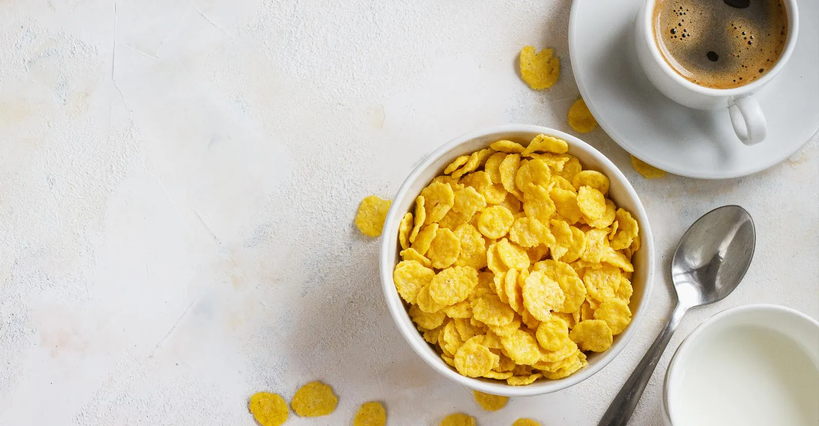 Bowl of cornflakes beside a cup of coffee and a jug of milk on a pale table