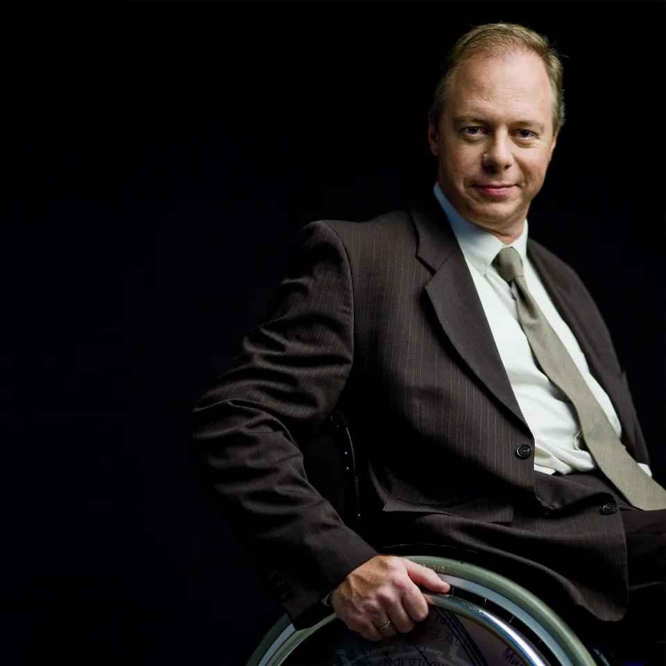 Portrait of a man in a pinstriped suit sitting in a wheelchair against a black background.