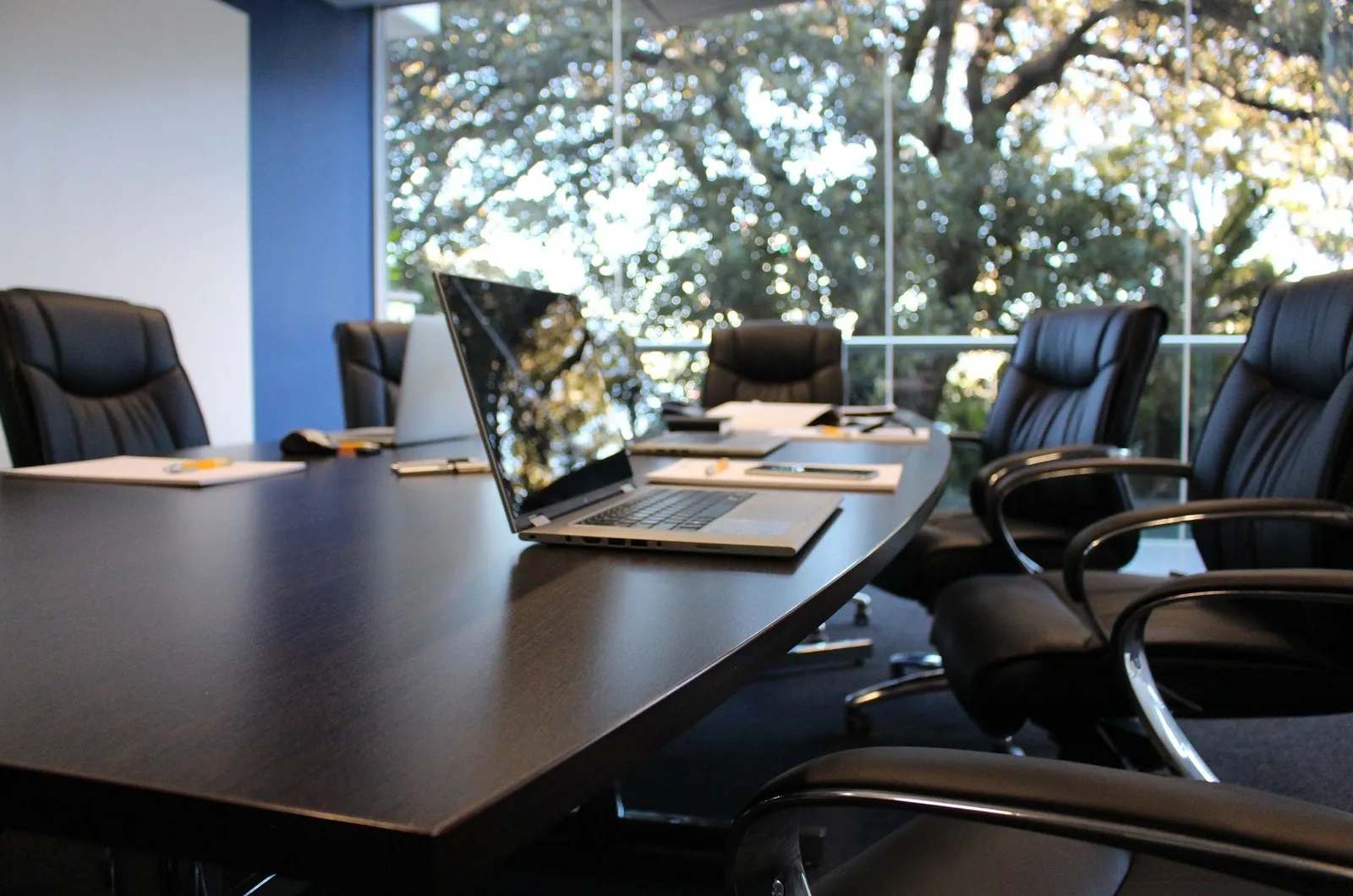 Empty boardroom with dark leather chairs around a long table, with laptops and notepads, and a window onto trees