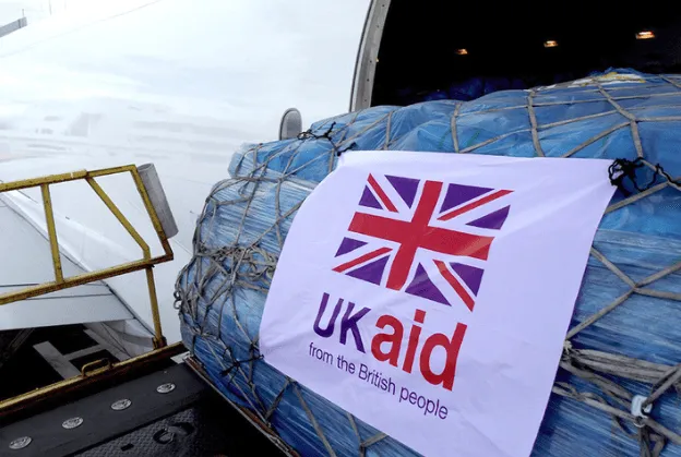 A banner with the UK flag and "UK aid from the British people" is attached to a cargo net on an aircraft.