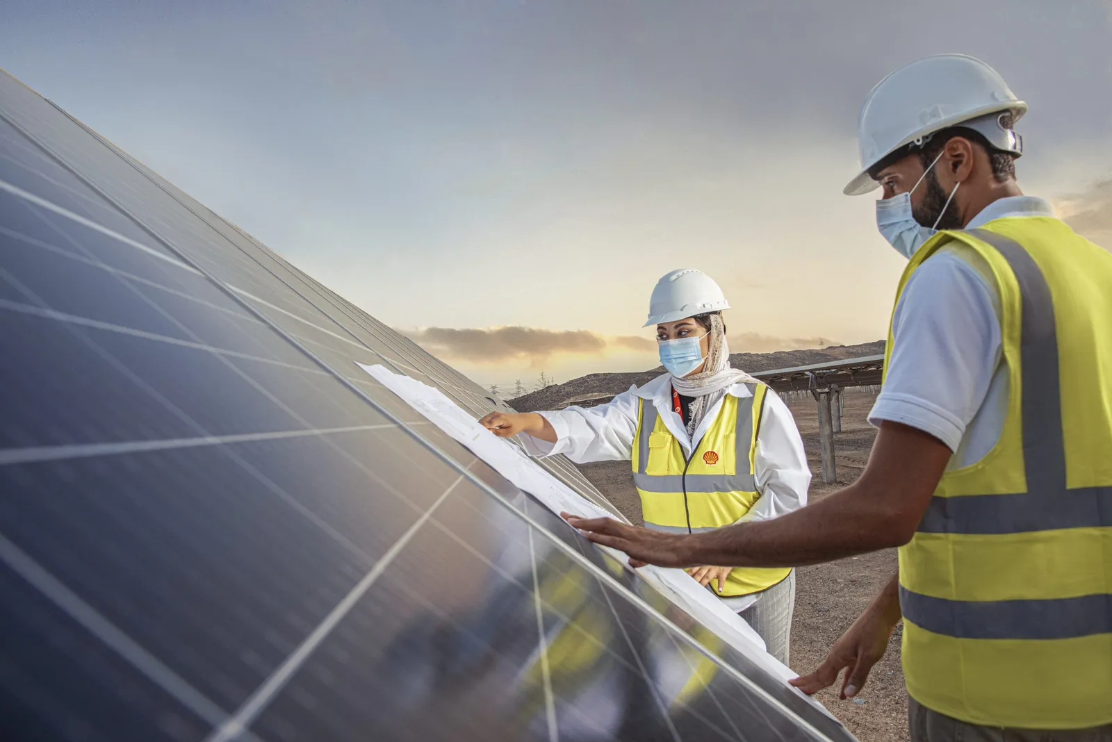 Two engineers in hard hats and high-visibility vests check plans on a solar panel array at sunset.