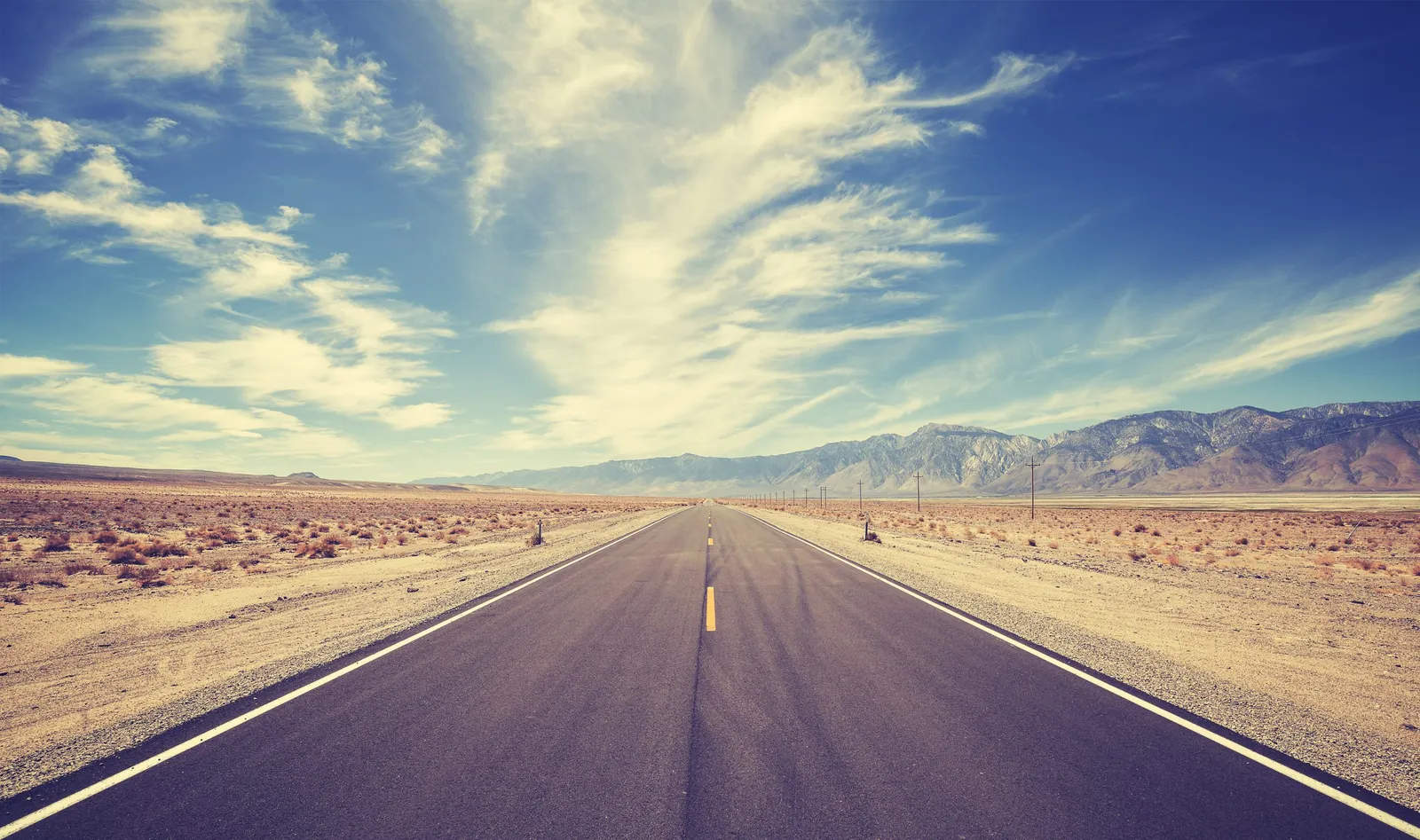 An empty road stretches across a desert towards mountains under a blue sky with wispy clouds.