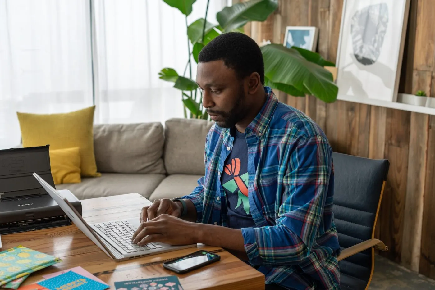 Man in a plaid shirt typing on a laptop at a wooden desk in a living room with a plant and a sofa behind him.