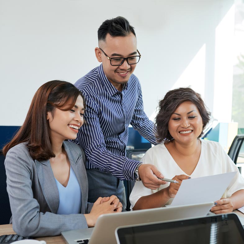 Three colleagues smiling over a document at a laptop, one standing behind the other two